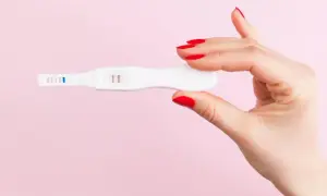 Dłoń z czerwonymi paznokciami trzyma biały test ciążowy z dwoma różowymi paskami.