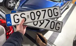 Dłoń trzyma polskie tablice rejestracyjne z numerem GS 0970G na tle niebieskiego samochodu.