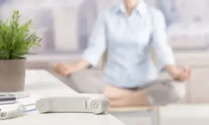 En person mediterar på kontoret. En telefonlur och en växt syns i förgrunden. En pausgympa på jobbet kan ge ny energi.