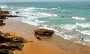 Skaliste marokańskie plaże z turkusową wodą i falami. Dwie osoby z deskami surfingowymi wchodzą do oceanu.