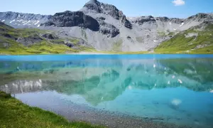 Ein klarer, blauer Bergsee, der **lai da rims**, spiegelt die majestätischen, grauen Berge und grünen Hänge wider.