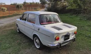 Vit Simca-bil parkerad på en gräsbevuxen stig, med olivträd och åkermark i bakgrunden.