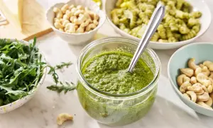 Świeże pesto z rukoli w słoiku, obok miseczki z orzechami nerkowca, rukolą i kawałkiem parmezanu.