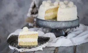 Una fetta di torta Raffaello, soffice e ricoperta di cocco, su un piatto decorato. Sullo sfondo, la torta intera su un'alzata.