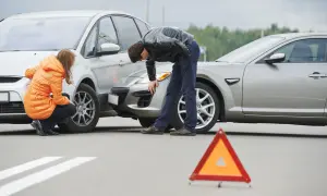 Incidente stradale con due auto danneggiate. Una donna in giacca arancione e un uomo esaminano i veicoli. Triangolo di segnalazione a terra. Nessun segno di omissione di soccorso.