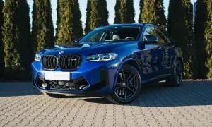 Błękitny suv BMW X4 M Competition błyszczy na tle zielonych drzew.