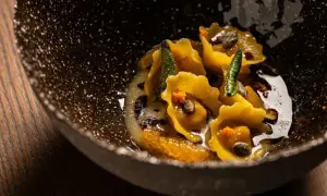 Tortelli di zucca dorati, guarniti con salvia croccante e arancia, in una ciotola scura e rustica.
