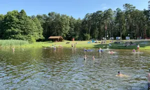 Słoneczny dzień nad jeziorem Szczutowo. Ludzie kąpią się, opalają na trawie i odpoczywają pod wiatą.