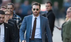 Mann im blauen Anzug, hellblauem Hemd und braunen Schuhen geht mit Sonnenbrille und Bart.