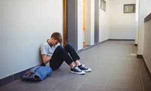 Chłopiec siedzi na korytarzu, oparty o ścianę, z ręką na czole. Obok leży plecak. Wygląda, jakby przegrał zabawę w krecika.