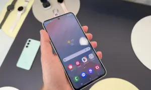 Ręka trzyma smartfon Samsung, pokazując ekran z pogodą i ikonami aplikacji. Obok leżą smartwatch i słuchawki. Czas przed ekranem Samsunga to chwila relaksu.