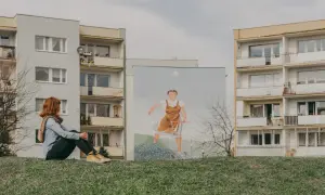 Dziewczyna z rudymi włosami siedzi na trawie, patrząc na mural z kobietą pchającą wózek na tle bloków na gdańskiej Zaspie.
