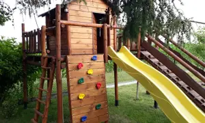 Drewniany plac zabaw z domkiem, zjeżdżalnią, ścianką wspinaczkową i drabinką.