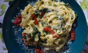 Pyszny makaron ze szpinakiem i suszonymi pomidorami, posypany parmezanem, na talerzu w kolorze głębokiego turkusu.