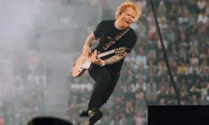 Ed Sheeran skacze na scenie, grając na gitarze, w tle tłum fanów.