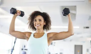 Femme souriante faisant des exercices avec haltères pour maigrir des bras. Elle est dans une salle de sport.