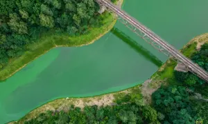 Widok z lotu ptaka na zieloną rzekę i most kolejowy. Woda ma intensywny, turkusowy kolor, a brzegi porasta bujna roślinność.