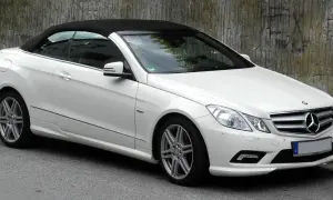 Biały kabriolet Mercedes W207 z czarnym dachem stoi na ulicy obok betonowej ściany.