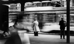 foto di strada: un uomo in impermeabile attende il tram, sfocato dal movimento. Altri passeggeri e la banca Commerzbank sullo sfondo.