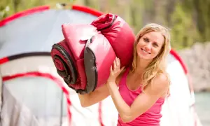 Lächelnde Frau trägt einen roten Schlafsack über der Schulter, bereit für Camping. Ob Daune oder Kunstfaser, er sieht bequem aus.