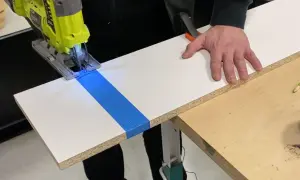 Une scie sauteuse coupe une planche de stratifié, guidée par du ruban bleu. La main d'un bricoleur stabilise la pièce.