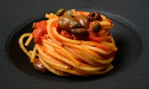 Ein kunstvoll aufgetürmter Teller Spaghetti Puttanesca, garniert mit Oliven und Kapern, auf schwarzem Teller.