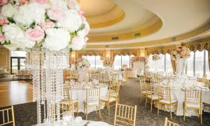 Elegancka sala weselna z okrągłymi stołami, złotymi krzesłami i bogatą dekoracją kwiatową. Stół państwa młodych ozdobiony jest wysokim bukietem róż i białych kwiatów.
