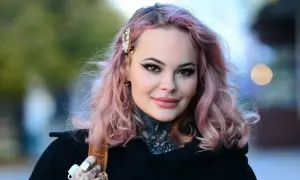 Monika Miller z różowymi włosami i tatuażami na szyi, mimo choroby, uśmiecha się do obiektywu.