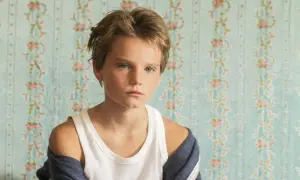 Un niño con cabello rubio y ojos azules, con una camiseta blanca y una chaqueta azul, parece un tomboy que es.