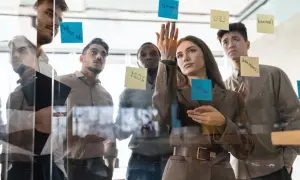 Team discute idee su lavagna di vetro, con post-it che indicano obiettivi e strategie per il **premio produzione**.