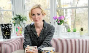 Jina Zachrisson sitter vid ett fönster med en mugg i handen, omgiven av blommor och konst.