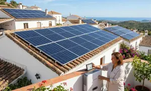 Mujer sonríe mientras revisa datos en tablet junto a inversor, con tejados llenos de paneles solares. ¡Un paso más para legalizar instalación fotovoltaica!