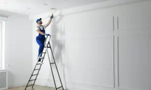 Mężczyzna w niebieskim kombinezonie i czapce maluje sufit wałkiem. Prace malarskie w jasnym, eleganckim wnętrzu.