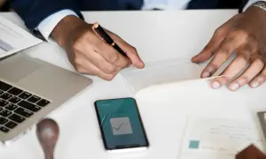 Uomo d'affari scrive su un libretto, con laptop e smartphone che mostrano grafici finanziari. Un esempio di come la categoria protetta art. 1 possa operare nel mondo del lavoro.