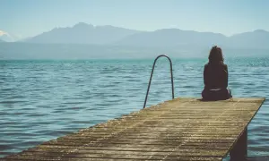 Mujer sentada en un muelle, contemplando el lago y las montañas. Pensamientos de frases de preocuparse por alguien flotan en el aire.