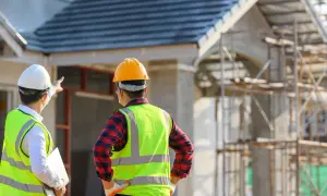Dwóch budowlańców w kaskach i kamizelkach odblaskowych omawia postęp prac na budowie. Widoczna tablica informacyjna budowy.