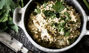 Cremiges Baba Ganoush, garniert mit Sesam und Petersilie, in einem rustikalen Topf. Daneben ein Messer und frische Kräuter.
