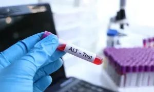 Ręka w niebieskiej rękawiczce trzyma probówkę z krwią do testu ALT (transaminaza GPT). W tle mikroskop i stojak z próbkami.