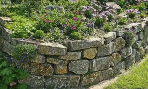 Eine **trockenmauer** sichert den **Hang** mit Natursteinen. Blumen und Grünpflanzen wachsen üppig auf und neben der stabilen **Hangbefestigung**.