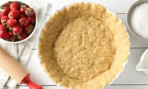 Gotowe ciasto na tartę z truskawkami czeka na nadzienie. Obok miska świeżych truskawek, cukier i dzbanuszek mleka.