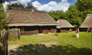 Drewniane chaty ze słomianymi dachami, trawiasty dziedziniec i ludzie.