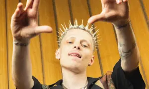 Raper Oki z blond irokezem, uniesionymi rękami i złotymi zębami na tle żółtej ściany.