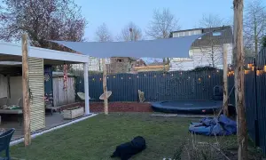Sonnensegel befestigen Ideen: Ein graues Sonnensegel spannt sich über einen Garten mit Trampolin und Sitzbereich. Holzpfosten dienen als Befestigungspunkte.