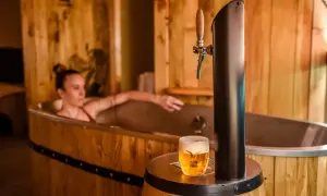 Entspannende Frau in einem Bierfass-Bad, daneben ein frisch gezapftes Pils. Ein einzigartiges Bier-Spa-Erlebnis in Pilsen.