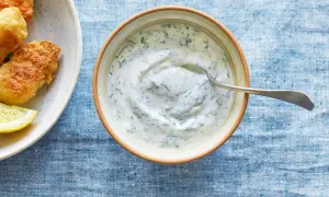 En skål med krämig kall sås till fisk, med dill och en sked. Bredvid syns friterad fisk och citronklyftor.