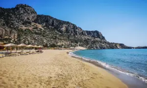 Piękna plaża Tsambika z piaszczystym brzegiem, turkusową wodą i skalistym zboczem w tle. Leżaki i parasole zapraszają do odpoczynku.