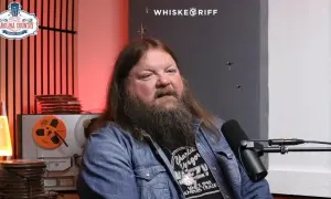 A man with a long beard and hair, wearing a denim jacket, speaks into a microphone. He's discussing Channing Wilson songs, possibly at a music festival.