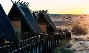 Hütten mit Strohdächern im Kgalagadi Transfrontier Park bei Sonnenuntergang. Ein Holzsteg führt zu den Unterkünften.