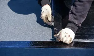 Manos enguantadas trabajando en la impermeabilización de tejados, cortando material negro sobre una superficie gris.