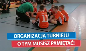 Trener w zielonej koszulce instruuje młodych piłkarzy w pomarańczowych strojach. To ważny moment przed kolejnym etapem mistrzostwa polski w chowanego.